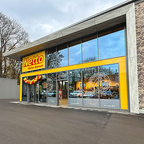 Der Eingang zu einem Netto Markt-Discount ist sichtbar, mit großen Fenstern und einer auffälligen, gelben Fassade. Bunte Luftballons schmücken den Eingang, und im Hintergrund sind Einkaufswagen und Kunden zu sehen. Der Himmel ist bewölkt.